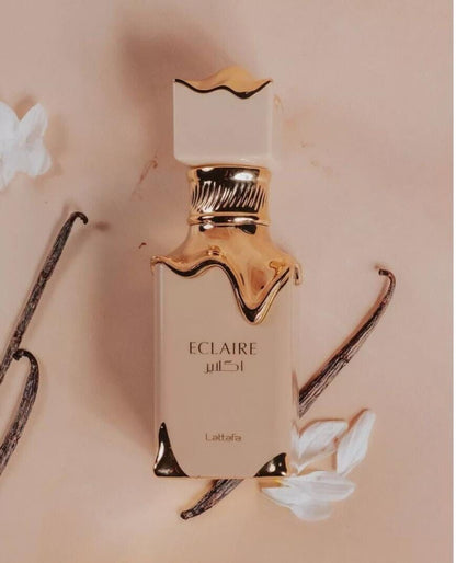 A bottle of Lattafa Eclaire EDP perfume with a beige label and gold accents, surrounded by vanilla pods and white flowers on a beige background.